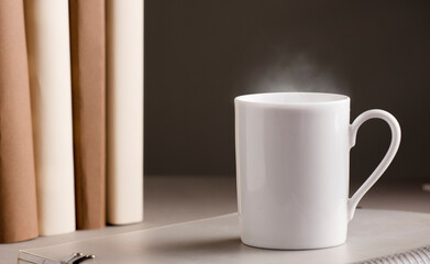 white cup of coffee with hot steam, on the desk with books, grey
 background