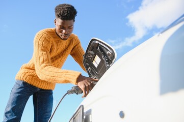 Electric cars, EV concept, eco friendly fuel. Portrait of young smiling black man, recharging his...
