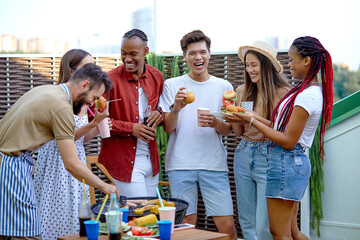 Happy young diverse friends eating burgers and drinking lemonade at barbecue patio party. Cheerful excited people having fun at bbq lunch. Youth lifestyle, summer and friendship concept. Portrait