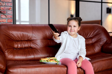 Fat Kid girl is going to eat hamburger and watch tv, sitting on sofa in living room after school,...