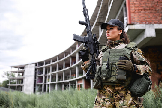 Military lady woman in tactical gear posing for photo in grass next to abandoned building. Beautiful female is Wearing green camo uniform holding assault rifle, looking at side, ready to shoot
