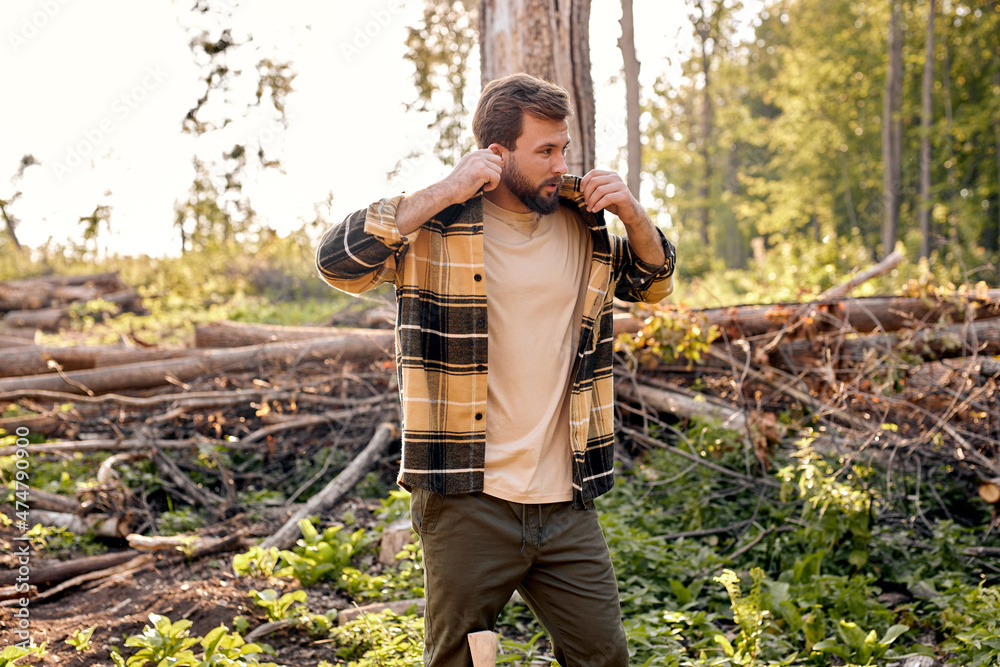 Wall mural Portrait of brutal bearded man lumberjack in casuak shirt is getting ready for cutting trees, wearing casual plaid shirt. european caucasian male is looking at side, trees in the background