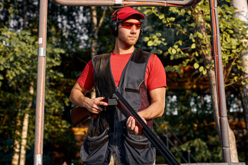Young caucasian man skeet shooting outdoors. shooting clay pigeon targets in outdoor range, stands looking at side in contemplation, wearing protective safety goggles and headset, in cap