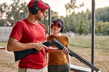 handsome confident man teach woman to recharge a rifle on shooting range outdoors, give instructions and have talk, wearing protective headset, spectacles and cap, holding rifle in hands. - obrazy, fototapety, plakaty