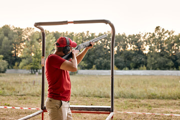 young caucasian man in goggles and headset aiming rifle at side, ready to shoot in an outdoor range. Skilled experienced man side view. Firearms for sports shooting, hobby. At summer evening