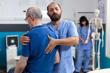 Orthopedic nurse stretching spinal cord and shoulder of elder patient with injury for physiotherapy. Chiropractor giving help to senior man with back pain for physical recovery. - obrazy, fototapety, plakaty
