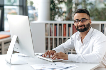 Fototapeta na wymiar Smart successful handsome Indian businessman, manager, office worker with beard and glasses. Business owner sitting at table in modern office, using computer, looking at camera, smiling