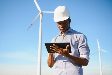 African engineer wearing white hard hat standing with digital tablet against wind turbine on sunny...