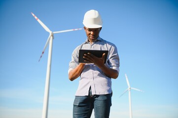 African engineer wearing white hard hat standing with digital tablet against wind turbine