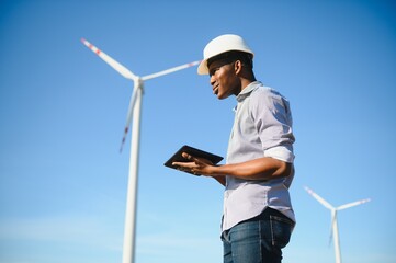African engineer wearing white hard hat standing with digital tablet against wind turbine