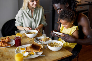 family having breakfast and using smartphone, parents with child in cozy kitchen, going to watch cartoons while eating delicious food, family time at weekends holidays at home. love concept