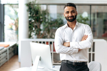 Fototapeta na wymiar Portrait of attractive, confident, smart, influential handsome Indian business leader, CEO, top manager, IT specialist, in a white shirt, stand near the table in the office, looks at the camera smiles
