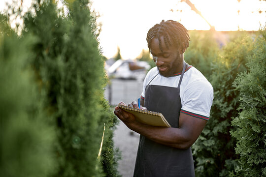 Focused black male writing notes at plants nursery, wearing uniform. man checking quality of plants and take notes on paper in garden greenhouse, Agricultural fresh organic concepts. at sunny day