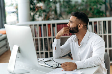 Pensive, intelligent, self-confident successful Indian businessman, top manager or ceo with beard sits at table, wears stylish shirt, looks to side, ponders business strategy or project.