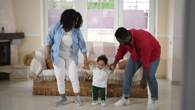 Wide Shot Happy African American Father Mother And Toddler Son Dancing In Living Room At Home. Positive Confident Millennial Couple Of Parents Having Fun With Cute Boy Indoors On Weekend