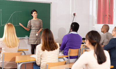 Concentrated Asian female teacher lecturing to adult students in auditorium, pointing at chalkboard and explaining function graph