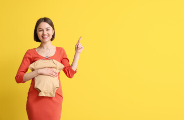 Young pregnant woman with baby bodysuit pointing at something on yellow background