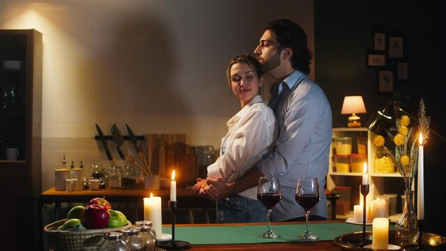 Man and woman dancing in cozy kitchen, romantic date dinner. Young husband and wife drinking red wine in evening. Loving couple, happy family relationships. Celebrating Valentine's Day together. 