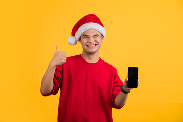 Smiling young Santa man wearing Christmas hat hold mobile phone with empty blank screen showing thumb up isolated on yellow background studio. New Year celebration holiday concep