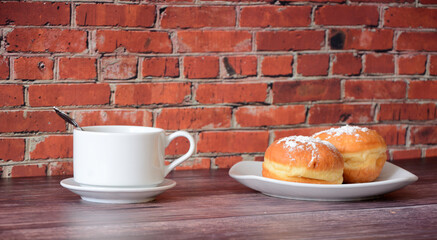 Two fresh donuts sprinkled with powdered sugar and a cup of coffee stand on a wooden table against the wall.