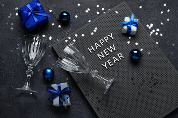 Board with text HAPPY NEW YEAR, gift boxes, glasses and Christmas decor on dark background