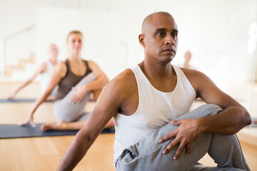 Fototapeta na wymiar Focused hispanic man doing yoga with group of people in fitness studio, sitting in twisting asana Matsyendrasana