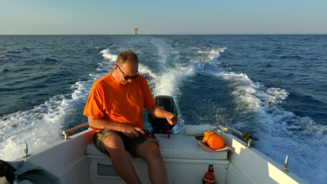 Man with sunglasses orange t-shirt and shorts on board of navigating motorboat holds steering tiller for direction while using smartphone. Slow-motion