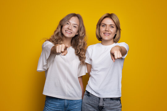 Portrait of a two women smiling and standing together while pointing at camera isolated on yellow background. Hey you