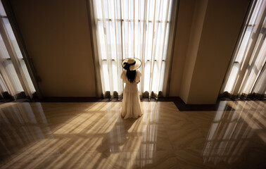 Back view of lonely young woman in white dress standing alone near the windows. Loneliness,...