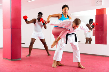 Asian woman exercising knee strike move with her trainer in kimono, African-american man boxing in front of mirror behind them.