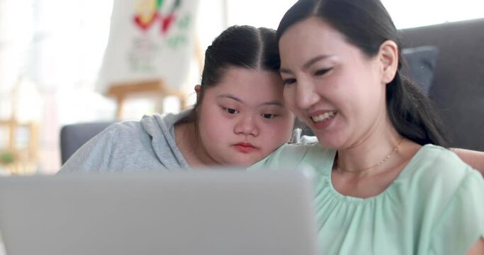 Cheerful loving ethnic mother and girl with down syndrome watching cartoon on netbook together and enjoying weekend at home