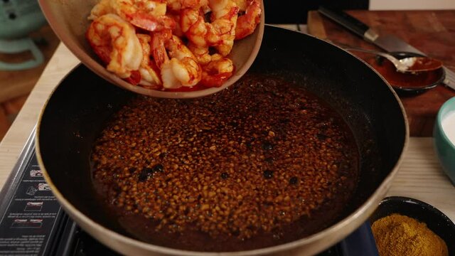 Slowly dropping tasty cooked shrimps into bubbling garlicky paprika hot sauce, coating the flavor onto the peeled shrimps, enrich the flavor and aroma of the seafood dish, close up shot.