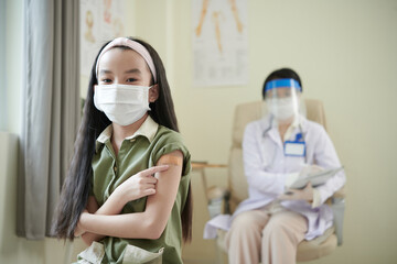 Portrait of girl in medical mask pointing at adhesive plaster over her covid-19 vaccine injection site
