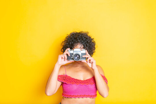 beautiful curly haired mexican young girl using old camera on yellow isolated background