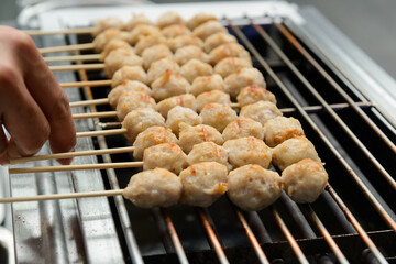 A merchant is flipping a skewer of pork meatballs with his hand to cook it on a smokeless electric grill. (selective focus)