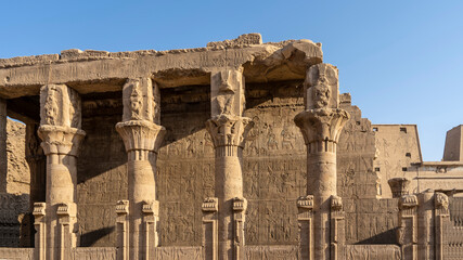 Obraz premium The dilapidated colonnade and wall in the ancient temple of Horus in Edfu are decorated with hieroglyphs, carved drawings. Capitals of columns of various shapes. Clear blue sky. A sunny day. Egypt