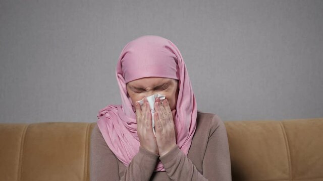 Young Muslim woman wearing pink-colored hijab catches cold and sneezes into paper napkin sitting on sofa in living room close view.