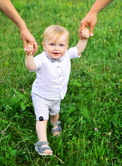 Parents hand and child first step. Baby playing in green grass. Child having fun on family picnic in summer garden. Portrait of a happy young family teaching baby to walk in the park.