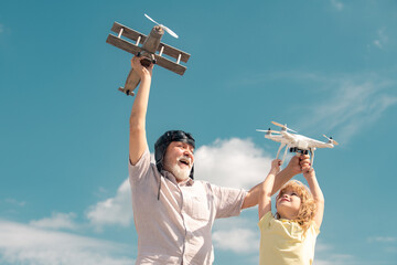 Old grandfather and young child grandson with toy jetpack plane and quadcopter drone against sky. Child pilot aviator with plane dreams of traveling.