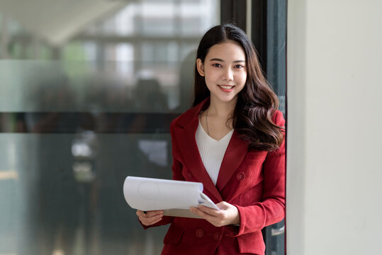 Happy young Asian businesswoman standing in a red suit holding and opening document in office. Looking at camera.
