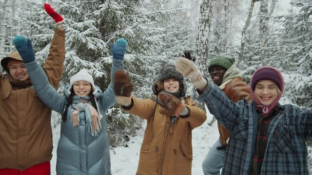 Group portrait of joyful multiethnic friends looking at camera, raising arms and yelling while posing in winter forest