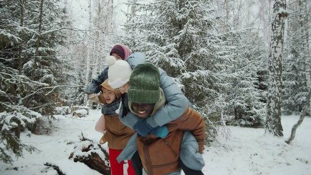 Young joyful friends doing piggyback ride in winter forest as girl filming video with smartphone
