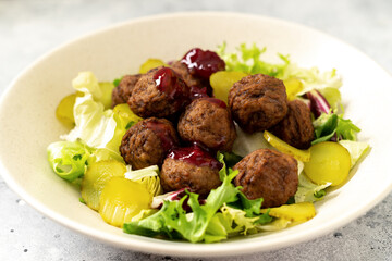 Swedish meatballs with salad, pickled cucumber and lingonberry jam in a bowl. Traditional Scandinavian dish in a ceramic plate on a light gray culinary background closeup