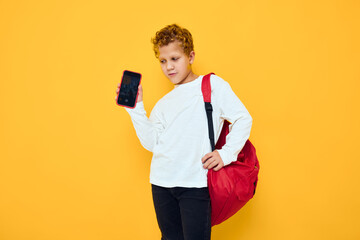smiling little boy in white sweatshirt mobile cell phone red backpack yellow background