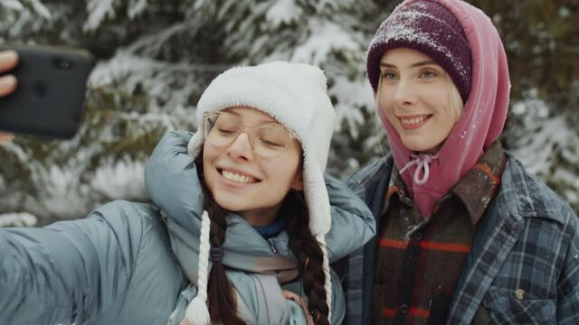 Two young beautiful women smiling and waving while taking selfie with mobile phone on walk in winter forest