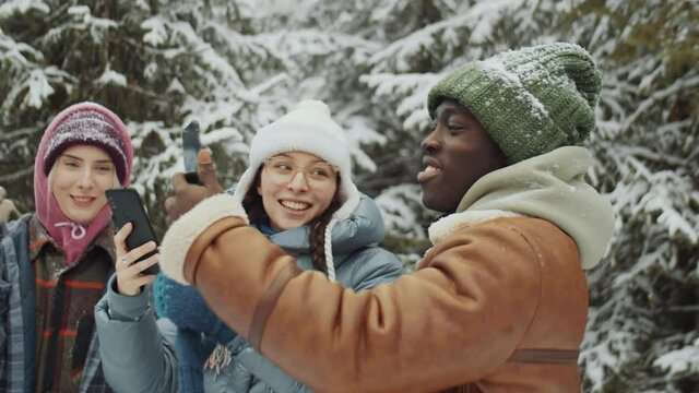 Two young Caucasian women and African American man talking and taking pictures of beautiful winter forest with phones