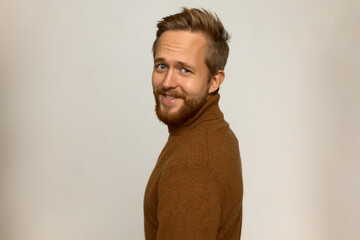 Obraz premium Handsome photogenic adult guy in good mood wearing cashmere turtleneck with stylish beard and haircut standing half-turned to camera, smiling against white wall with copy space for your content