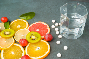 Supplements or healthy natural food? Scattered white pills vitamins, Sliced citrus fruits, orange, lemon, kiwi, grapes, glass with water on a stone gray background