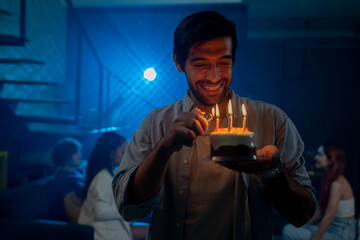 Portrait of smiling young and handsome man lighting candles on a birthday cake with friends...