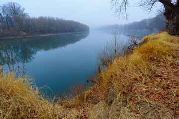 wild natural landscape, late autumn season, river, bare branches of trees without leaves, cloudy weather with haze, forest with silhouettes of trees